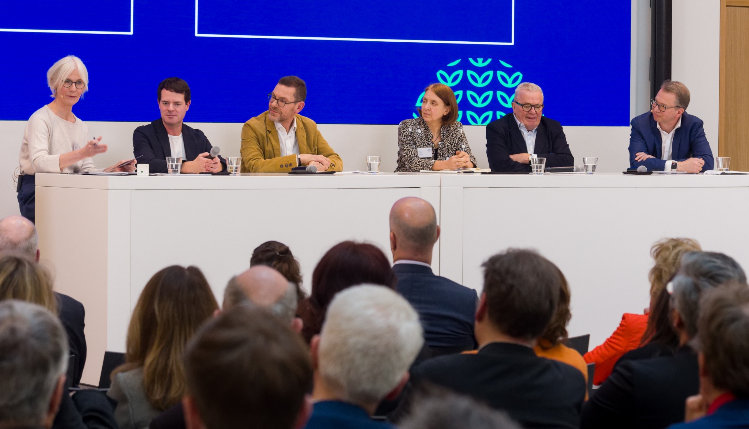 Zu sehen sind sechs Personen, die nebeneinander auf einem erhöhten Podium sitzen.