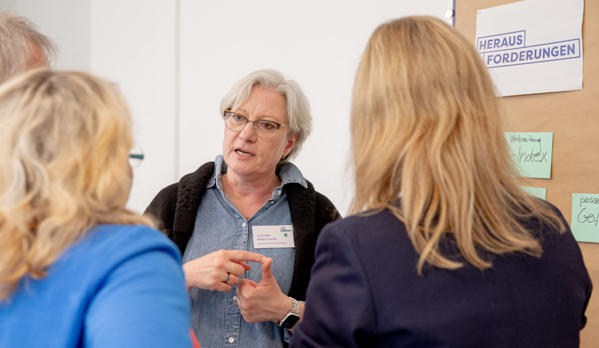 Auf dem Foto befindet sich eine Frau im Gespräch mit drei anderen Personen. Im Hintergrund ist eine Stellwand auf der das Wort "Herausforderungen" zu lesen ist.