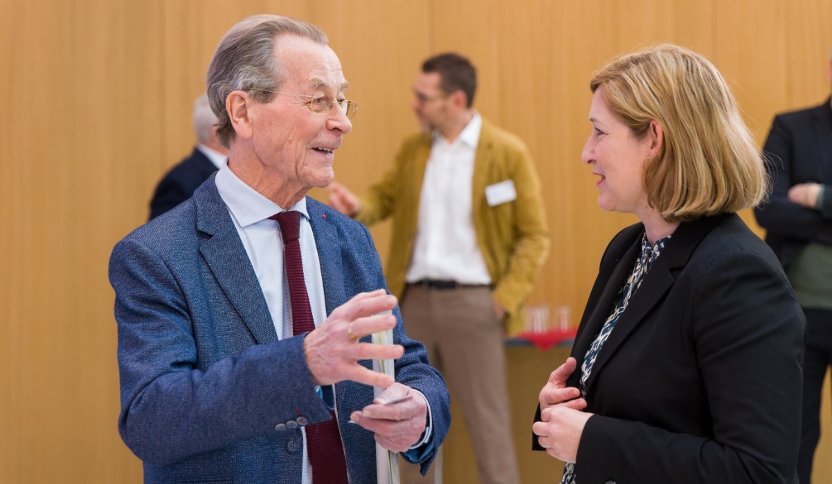 Zu sehen sind ein Mann im Anzug und eine Frau mit Blazer. Sie führen ein angeregtes Gespräch.