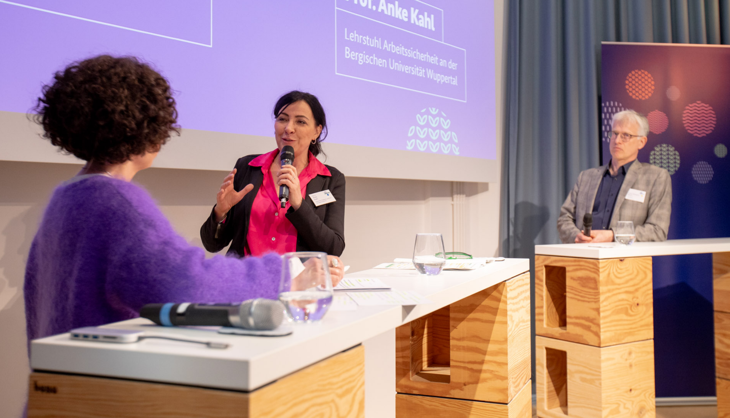 Zu sehen sind drei Personen im Dialog auf dem Podium.