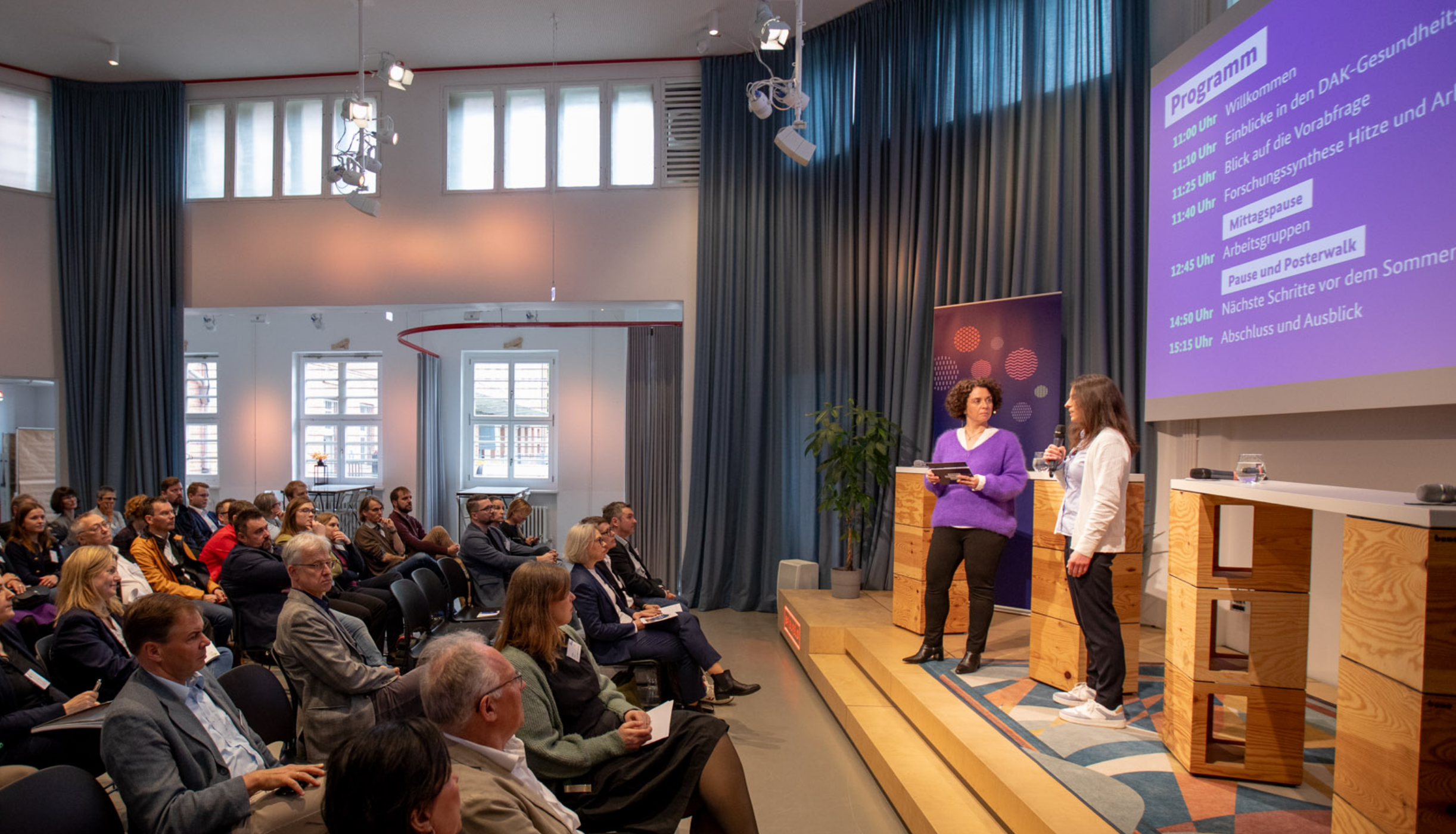 Zu sehen ist ein Foto der Dialogveranstaltung. Auf der rechten Seite stehen zwei Frauen auf einer Bühne. Gegenüber sitzen mehrere Personen im Publikum.