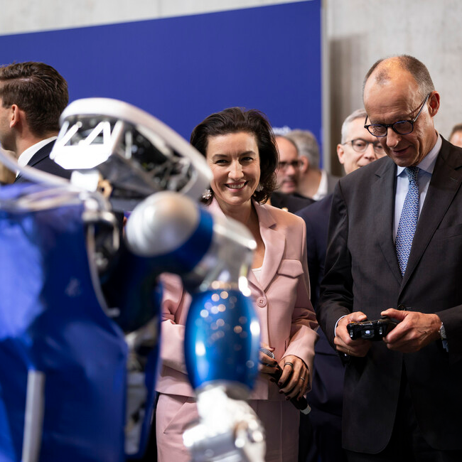 An einem Ausstellungsstand bedient Bundeskanzler Friedrich Merz einen Roboter mit einem Controller