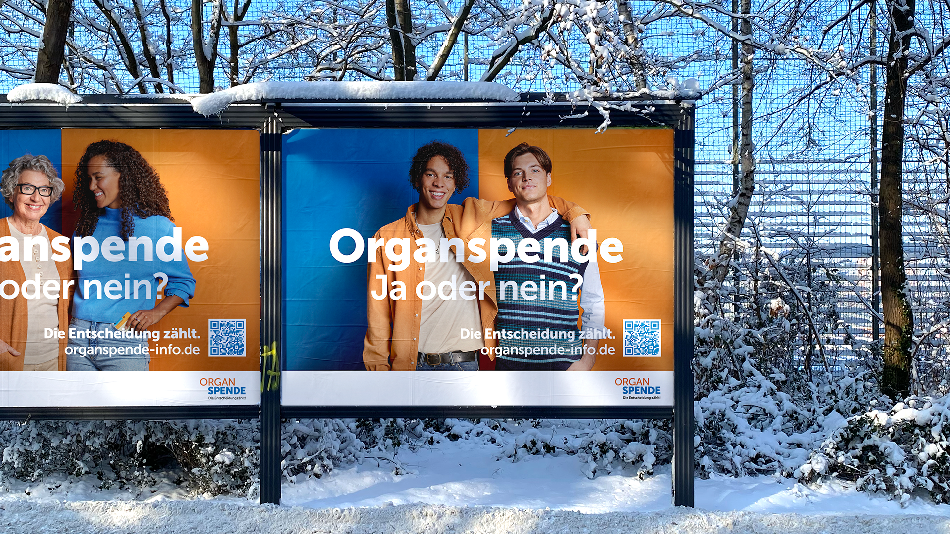 Gro&szlig;fl&auml;chenplakete der Kampagne im Schnee. Je zwei M&auml;nner und zwei Fraune halten sich im Arm vor abwechselnd orangem und blauen Hintergrund.