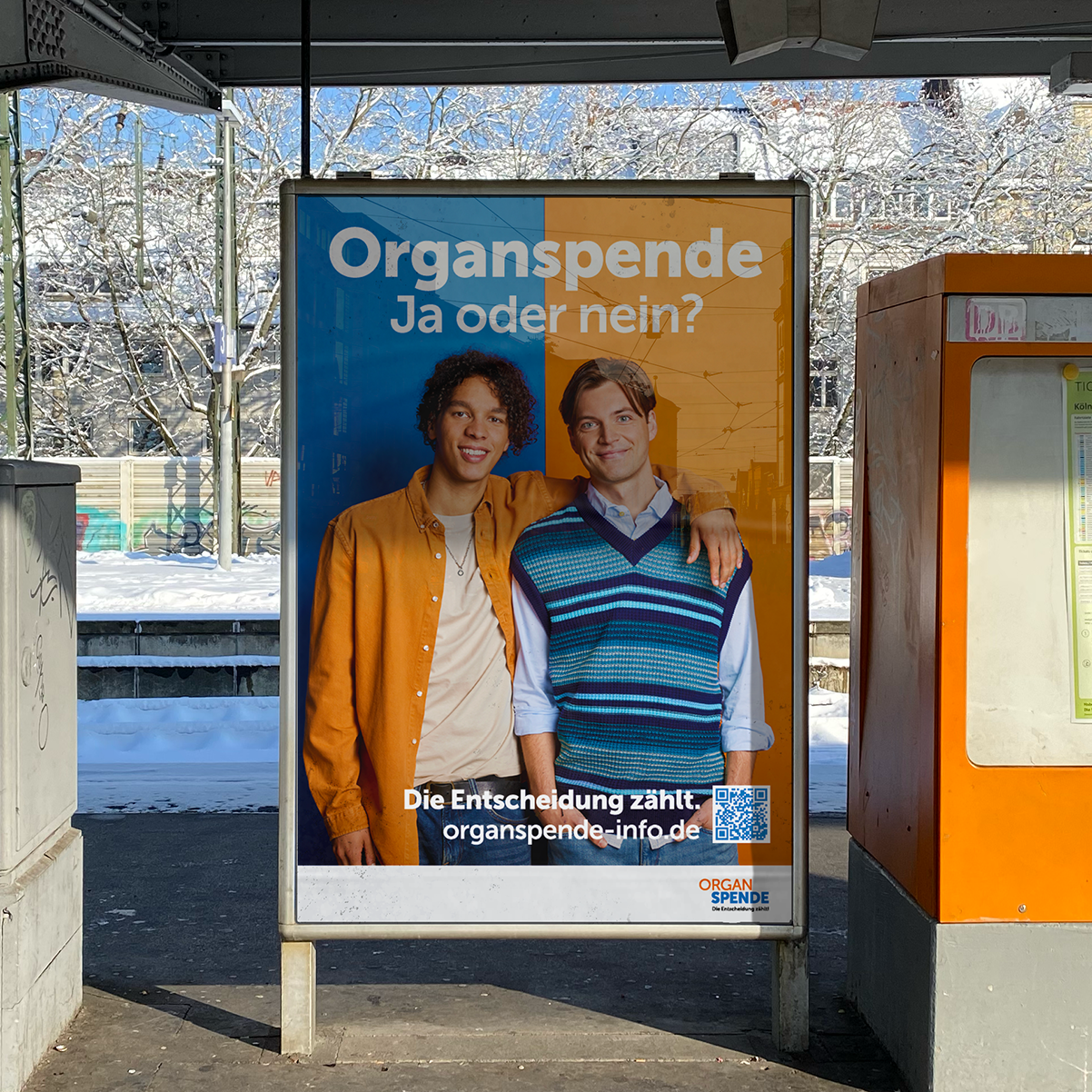 Gro&szlig;fl&auml;chenplakat der Kampagne an einem Bahnhof. Zwei M&auml;nner halten sich im Arm vor abwechelnd blauen und orangenem Hintergrund.