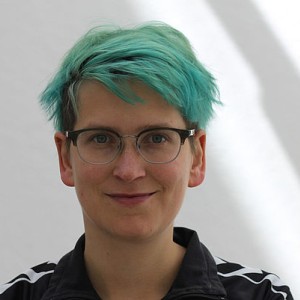 Portraitfoto von Eva Horn. Sie trägt eine Brille, türkis-grün gefärbte kurze Haare und lächelt in die Kamera.