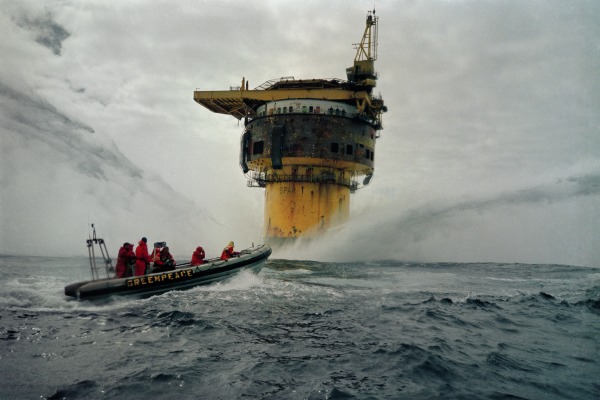Ein kleines Boot von Greenpeace mit mehreren Menschen an Board vor einer &Ouml;lplattform auf dem offenen Meer. Es wird sehr viel Wasser aus der linken und rechten Richtung zur &Ouml;lplattform gespritzt.