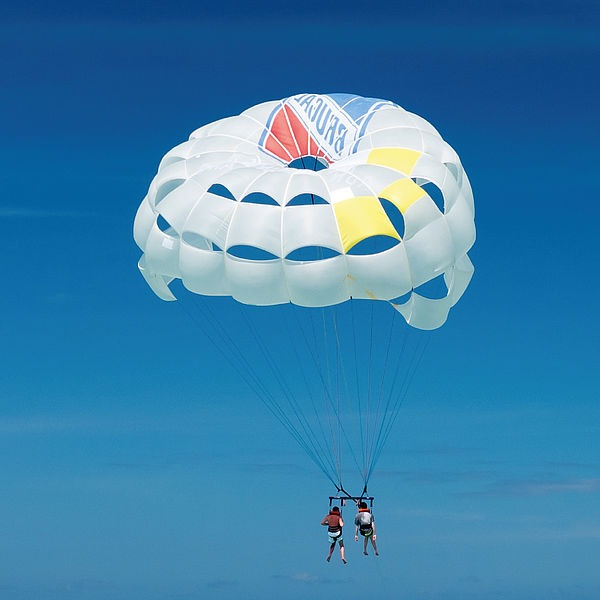 Zwei Menschen fliegen mit einem Fallschirm. Hinter ihnen sieht man den blauen Himmel. In diesem Bildausschnitt ist nicht zu sehen, dass die Menschen nur knapp &uuml;ber dem Meer &uuml;ber einem Boot gleiten.