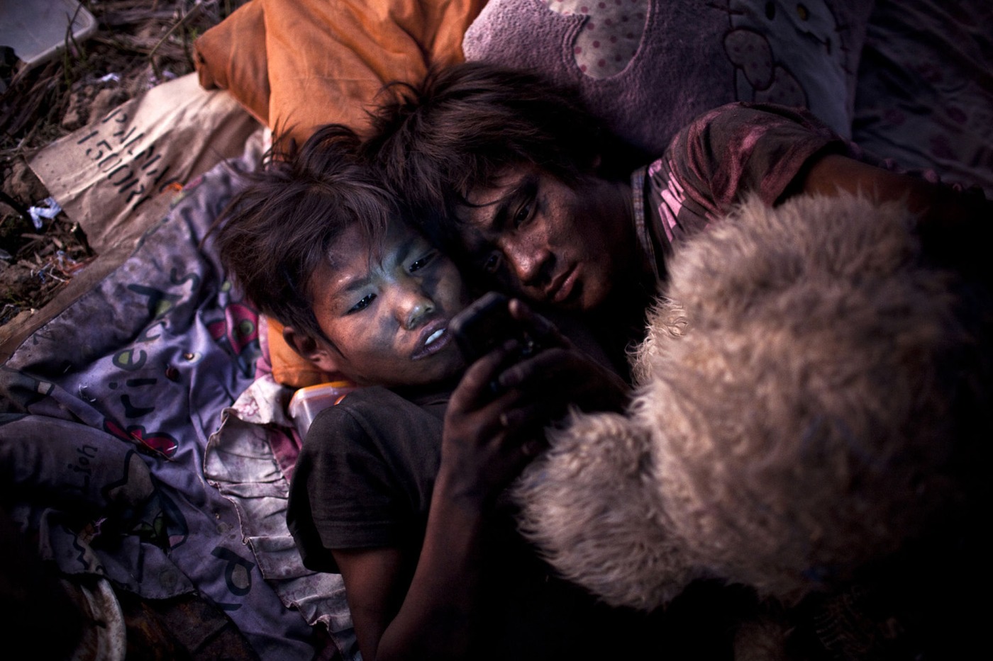 Zwei Kinder mit schmutzigen Gesichtern schauen zu zweit auf ein Smartphone-Display. Sie sitzen zwischen M&uuml;ll auf einer Deponie in Thailand.