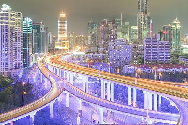 Panorama der Gro&szlig;stadt Shanghai mit Blick auf eine mehrspurige Autobahn, die zwischen den Wolkenkratzern durch f&uuml;hrt. Die Autobahn sowie einige Wolkenkratzer sind in lila-Farben erleuchtet.